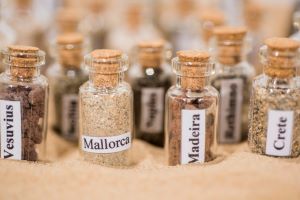 Viele kleine Apothekerfläschen stehen im Sand, mit verschiedenem Sand Inhalt, beschriftet mit orten wie Madeira und Mallorca