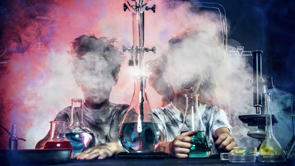 Deux enfants sont assis à une table, devant eux des bocaux de laboratoire munis de tubes et remplis de liquides colorés. Partout, ils se couvrent le visage