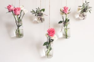Verschiedene Gläser und Flaschen gefüllt mit Wasser und einzelnen rosa Blumen, die an Schnüren an der Wand befestigt sind