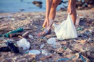 Ein Strandausschnitt übersät mit Plastikmüll, im Hintergrund eine Person, die sich bückt, um Müll aufzuheben, in der anderen Hand eine Tüte. Man sieht nur Beine und Hände