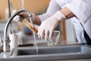 Ein Wasserhahn in einem Labor, zwei Hande mit Handschuhen und Kittelärmeln, die einen Erlenmeyerkolben unter fließendem Wasser mit einer Bürste reinigen