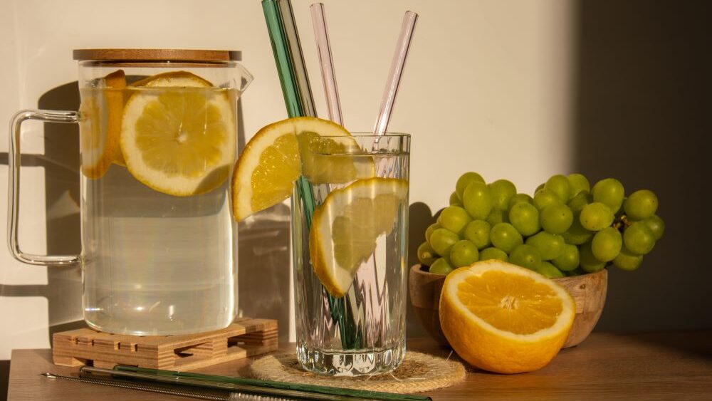 Sur une table se trouve un pichet contenant de la limonade et des tranches de citron, à côté un verre avec une tranche de citron et des pailles en verre sur le bord. Autour, une demi-orange et une grappe de raisins verts, encore plus de pailles en verre et un pinceau.