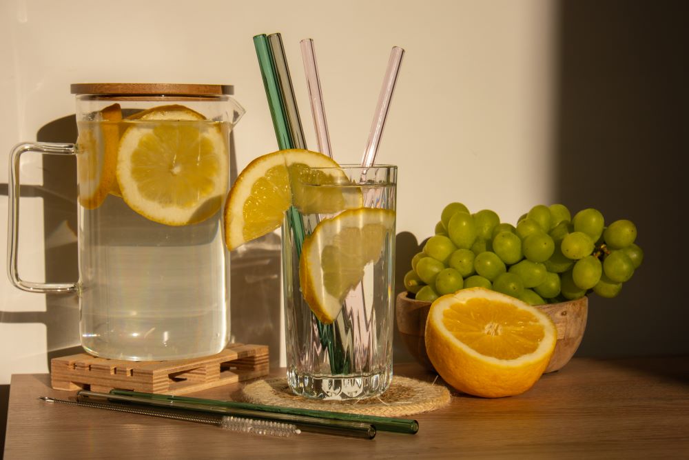 Sur une table se trouve un pichet contenant de la limonade et des tranches de citron, à côté un verre avec une tranche de citron et des pailles en verre sur le bord. Autour, une demi-orange et une grappe de raisins verts, encore plus de pailles en verre et un pinceau.
