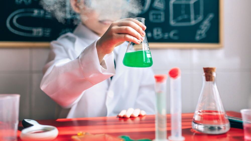 Un enfant en blouse de laboratoire devant un tableau noir avec des formules tient dans sa main droite une fiole d'Erlenmeyer contenant un liquide vert qui fume. Devant l'enfant, une table avec des verres et du matériel de laboratoire.