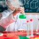 Un enfant en blouse de laboratoire devant un tableau noir avec des formules tient dans sa main droite une fiole d'Erlenmeyer contenant un liquide vert qui fume. Devant l'enfant, une table avec des verres et du matériel de laboratoire.