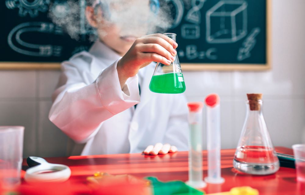 Un enfant en blouse de laboratoire devant un tableau noir avec des formules tient dans sa main droite une fiole d'Erlenmeyer contenant un liquide vert qui fume. Devant l'enfant, une table avec des verres et du matériel de laboratoire.