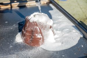 Un ramequin brun sur une assiette noire. Un liquide transparent provenant d'une seringue est versé goutte à goutte dans le ramequin par le haut et de la mousse blanche s'élève du récipient brun