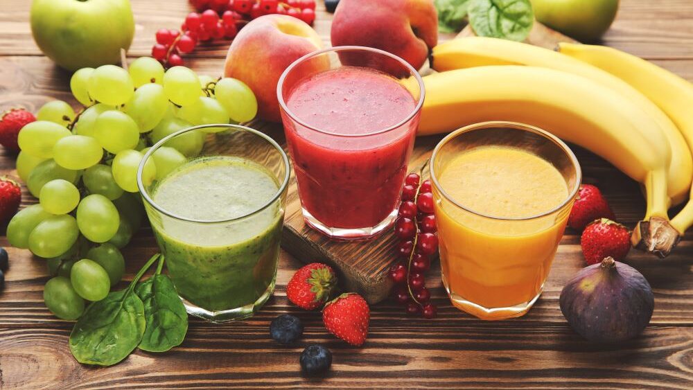 Trois verres contenant, de gauche à droite, un smoothie vert, rose et orange, de haut en bas sur une surface en bois avec différents fruits.