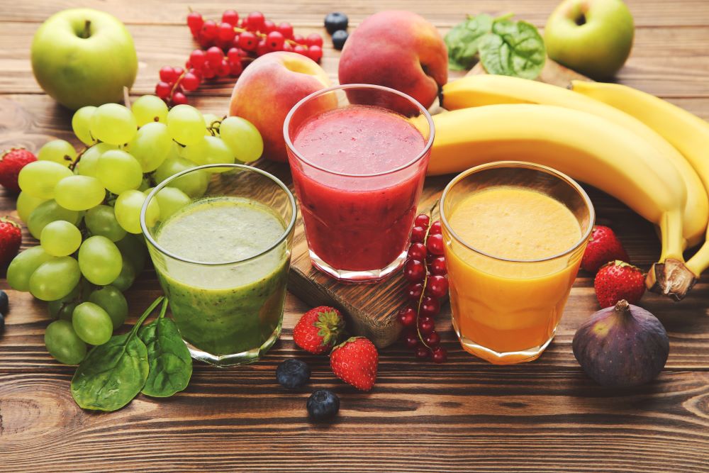 Trois verres contenant, de gauche à droite, un smoothie vert, rose et orange, de haut en bas sur une surface en bois avec différents fruits.